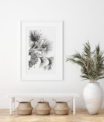 Framed print of palm leaves on white wall with pot plant on the right hand side.